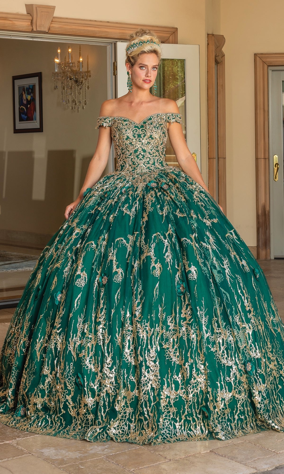 A woman stands indoors wearing the Quinceanera Dress 1742 by Dancing Queen, an off-the-shoulder emerald green ball gown with a sweetheart neckline, gold embroidery, and a matching headpiece.